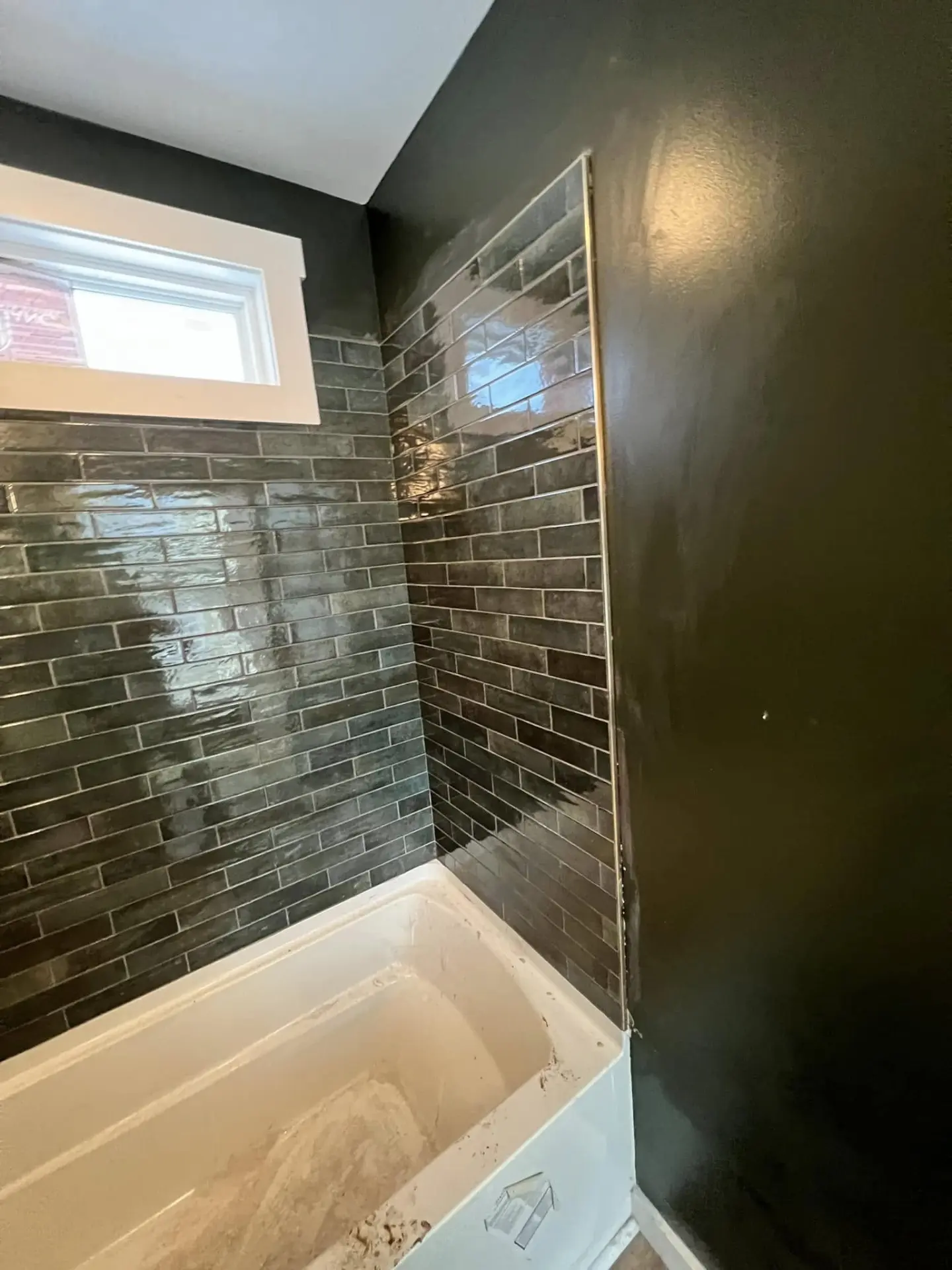 Finished shower with glossy dark green subway tile, gold accent trim and a tub