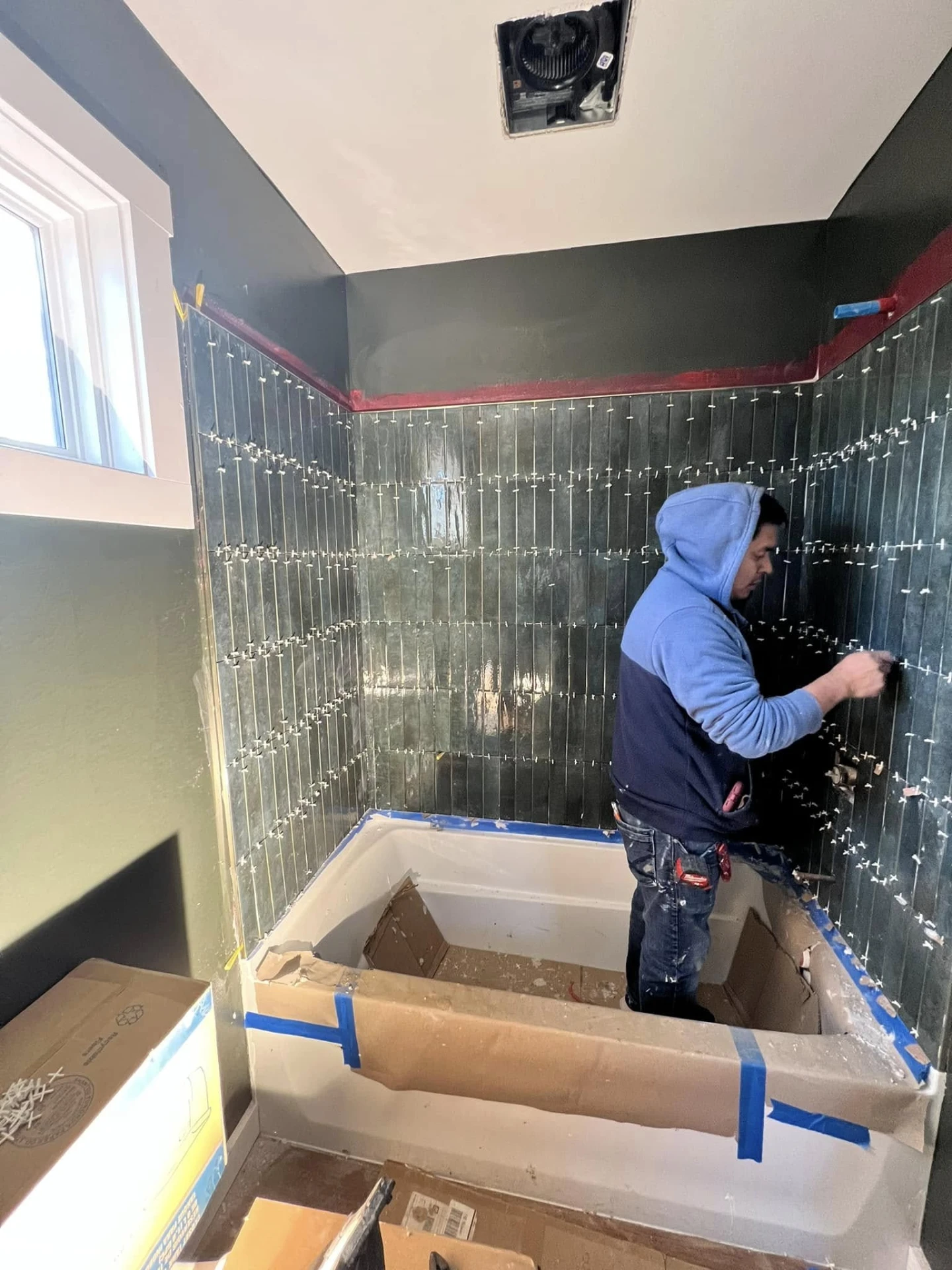 Tile installer setting dark green subway tile on a shower wall around a tub